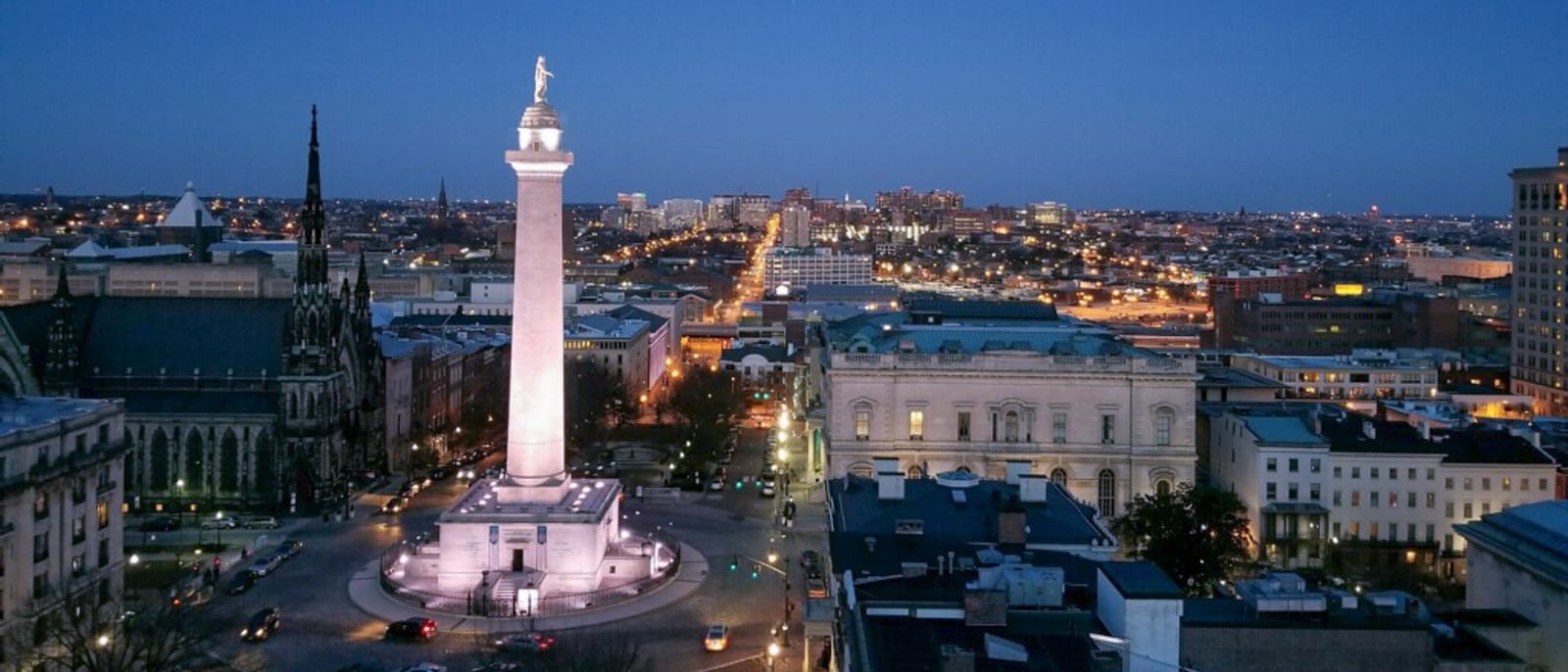 Aerial view of Baltimore's Washington Monument in Mount Vernon Place at dusk, the lit column rising above rooftops with the city's lights beyond.